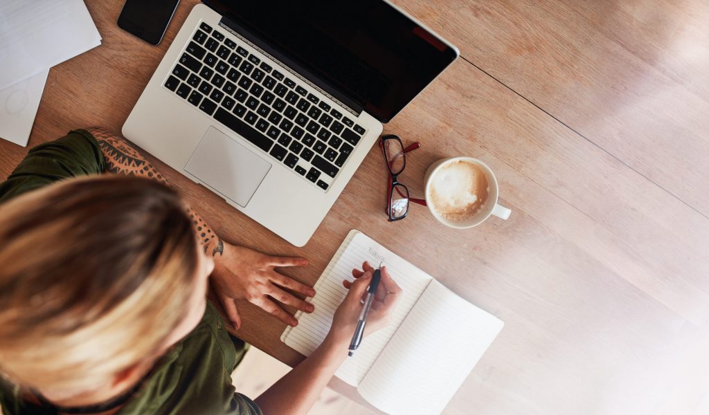 Top view shot of woman sitting at table with laptop and coffee writing on notebook. Female making to do list on diary.